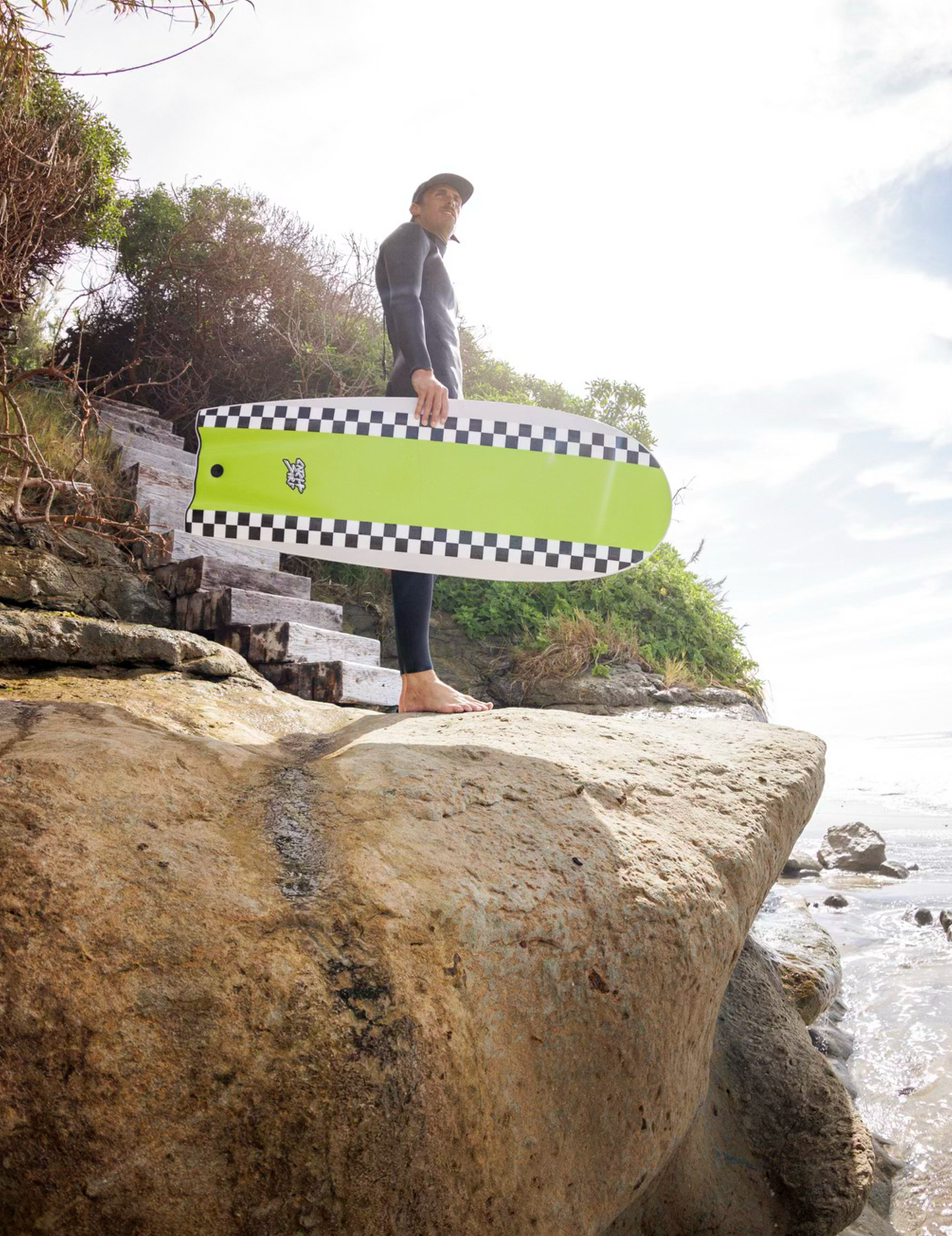 Person holding a green and black checkered surfboard on a rocky beach