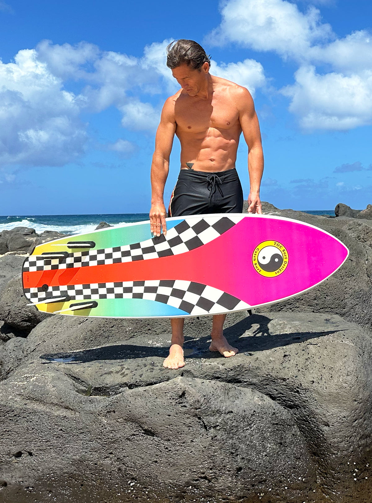 Man holding a colorful surfboard on rocks with ocean
